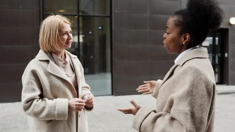 2 women talking outside a corporate building. They are wearing warm beige coats.